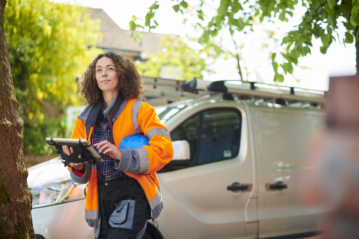 A woman with curly brown hair, wearing an orange high-visibility jacket, a dark shirt, and dark pants, stands outdoors looking slightly to her left while holding a black tablet. She has a blue hard hat tucked under her arm. A white work van with roof racks is parked behind her, and trees and a building are visible in the blurred background.