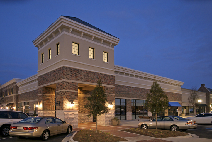 The image shows the exterior of a brick building at sunset, featuring several storefronts including retail and sports-related shops. There is drive-up parking available in front of the building.
